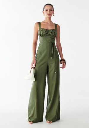 Olivengrøn jumpsuit med firkantet halsudskæring, rynket bodice, vide ben og bælte i taljen. Tilbehør med hvid håndtaske.
