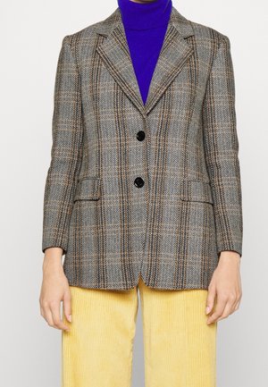 Kvinde iført en brun mønstret blazer over en blå rullekrave og gule ribbukser mod en ensfarvet baggrund.