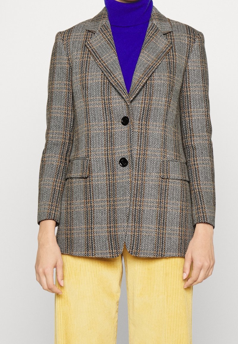 Femme portant un blazer marron à motif par-dessus un col roulé bleu et un pantalon en velours côtelé jaune sur un fond uni.