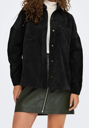 Kvinde iført sort kordfløjlsjakke med knapper, hvid rullekrave og mørkegrøn lædermininederdel med lynlås foran.