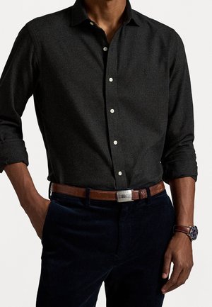 Homme portant une chemise à manches longues gris foncé boutonnée avec les manches retroussées, une ceinture en cuir marron, un pantalon bleu marine et une montre au poignet gauche.