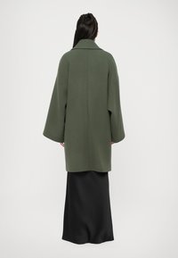 Oversized green wool coat with wide sleeves, featuring a collar and straight cut, worn over a black floor-length skirt.