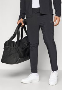 Zwarte sportbroek met een taps toelopende pasvorm, elastische tailleband en logo-details, gecombineerd met een zwarte sporttas en witte sneakers.