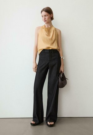 Vrouw draagt een mouwloze mosterdgele top, zwarte broek met hoge taille en wijd uitlopende pijpen, zwarte sandalen, zilveren armband en houdt een kleine gevlochten handtas vast.