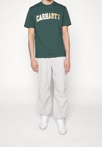 Zielona bawełniana koszulka z złotym logo "CARHARTT", połączona z jasnoszarymi spodniami cargo i białymi sneakersami, charakteryzująca się luźnym krojem i elastycznymi mankietami.