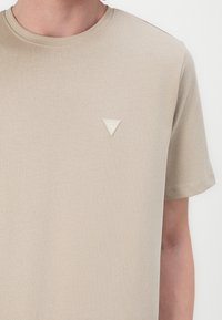T-shirt en coton beige avec un col rond et des manches courtes. Patch logo triangulaire iconique dans une teinte plus claire sur la poitrine gauche.