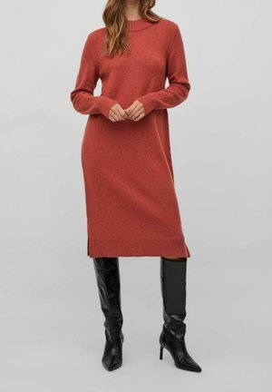 Žena v midi šatech s dlouhým rukávem, pletených v rezavé barvě, doplněná černými kozačkami na podpatku až po kolena, na jednobarevném šedém pozadí.