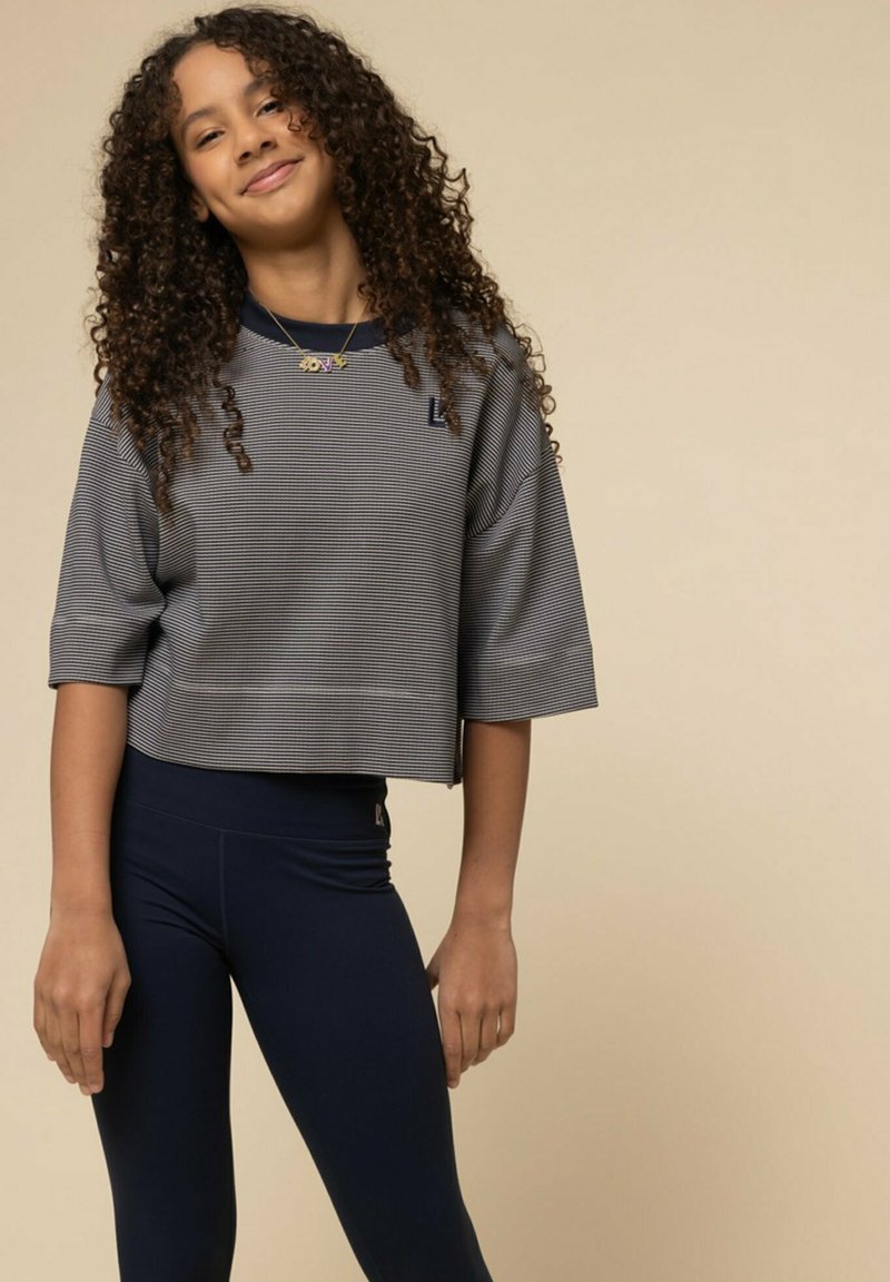 Grijs gestreept cropped top met brede mouwen, gecombineerd met marineblauwe leggings. Opvallende details zijn het logo op de top en de gladde stoftextuur.