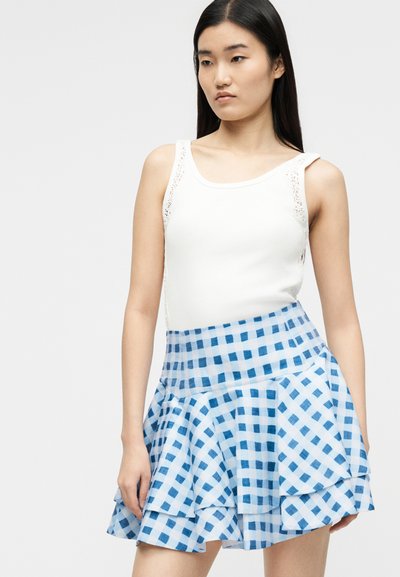 Νεαρή γυναίκα που φοράει λευκό αμάνικο τοπ με δαντέλα στα πλάγια και φούστα με μπλε-άσπρο καρό σε στρώσεις, στέκεται μπροστά σε απλό φόντο.