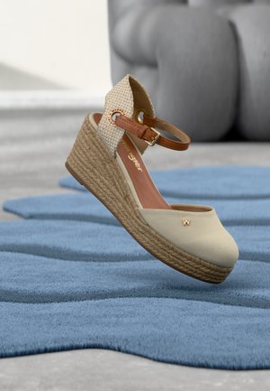 Béžový sandál na klínu s tkanou platformou, páskem kolem kotníku a detailem s logem "W", umístěný nad modrým texturovaným kobercem a na pozadí šedého nábytku.