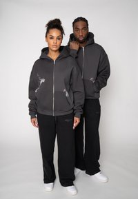 Graue Kapuzenpullover mit Reißverschluss und Stickerei; getragen zu schwarzen Sweatpants. Die Modelle stehen zusammen vor einem neutralen Hintergrund. Weiße Sneaker vervollständigen den Look.