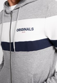 Sudadera con capucha gris con cremallera y rayas horizontales en azul marino y blanco. Presenta la palabra "ORIGINALS" en texto azul marino en negrita en el pecho. Capucha ajustable.