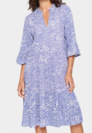 Vrouw draagt een losse jurk tot op de knie met blauw-witte abstracte bloemenpatronen en driekwartmouwen met klokvorm.