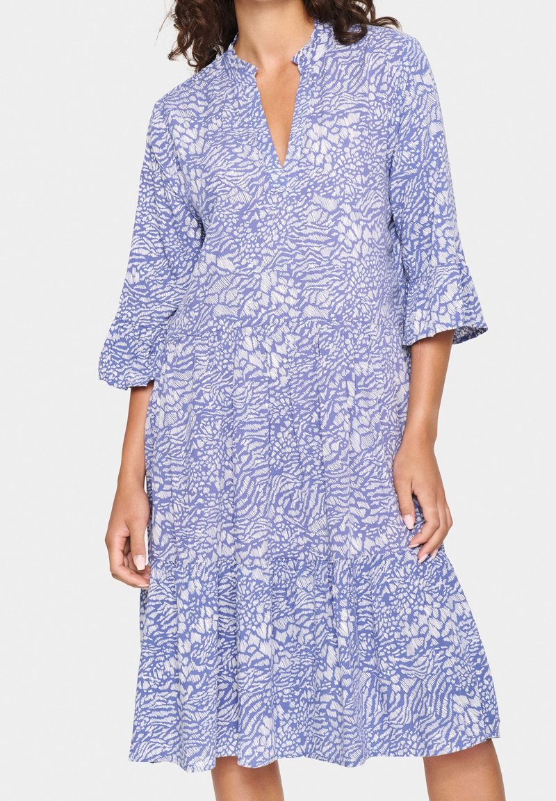 Femme portant une robe ample longueur genoux avec des motifs floraux abstraits bleus et blancs et des manches cloche trois-quarts.