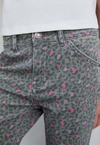 Jeans de mezclilla con base verde y un estampado abstracto de leopardo en rosa y púrpura claro. Incluye bolsillos delanteros y un cierre de botón plateado.