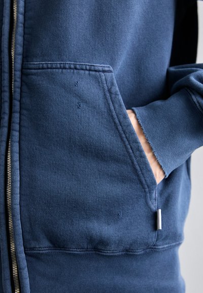 Mørkeblå zip-up hettegenser med teksturert stoff, med en frontlomme og slitte mansjetter. Glidelåsdetalj langs siden.