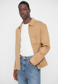 Tan wool jacket with a zip front, collar, and one chest pocket, worn over a white shirt and blue high-waisted jeans. Smooth texture.