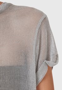 Gros plan d'une personne portant un haut en maille gris clair, tricoté lâchement, à manches courtes, par-dessus un sous-vêtement noir.