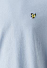 Jasnoniebieska bawełniana koszulka z małym czarnym i żółtym logo orła na piersi, o gładkiej fakturze i casualowym wzorze.