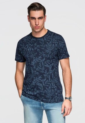 Mann trägt ein marineblaues Kurzarmhemd mit hellblauen Blattmustern und hellen Jeans, steht mit einer Hand in der Tasche.