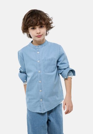 Jeune personne aux cheveux bouclés bruns portant une chemise bleu clair boutonnée avec les manches retroussées et un jean bleu, debout devant un fond blanc.