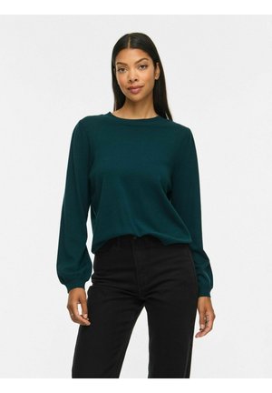 Vrouw die een donkergroene trui met lange mouwen en een zwarte broek draagt, staand tegen een effen witte achtergrond.