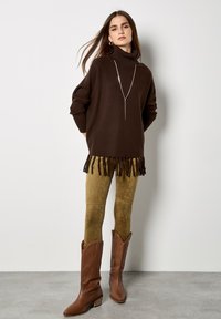 Brązowy sweter z golfem i frędzlami na dole, w połączeniu z oliwkowymi legginsami imitującymi zamsz oraz brązowymi butami w stylu western. Długa srebrna naszyjnik jako akcent.