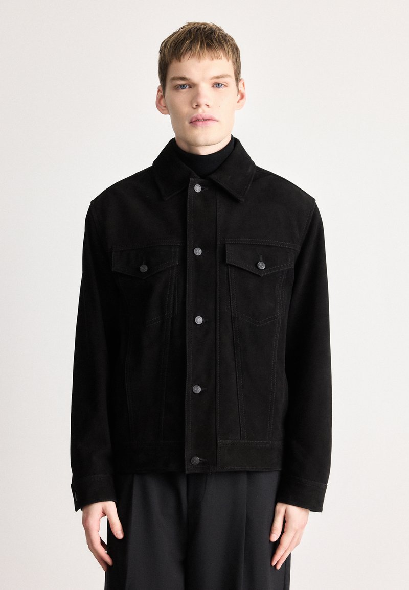 Melna žāvēta zamšāda jaka ar apkakli, pogu aizdari, divām krūšu kabatām un kontrastējošām šuvju detaļām. Gluda tekstūra ar strukturētu piegriezumu.