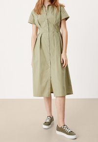 Světle zelené košilové šaty s krátkými rukávy a sklady, s knoflíkovým zapínáním vpředu; v kombinaci s olivově zelenými teniskami a bílými podrážkami.