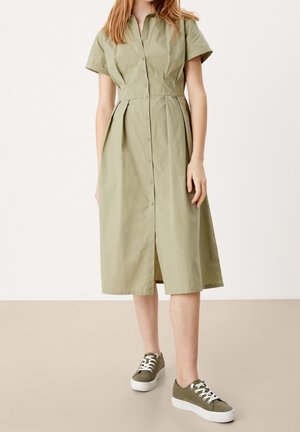 Abito camicia verde chiaro con colletto, bottoni frontali, maniche corte e gonna plissettata; abbinato a sneakers verde oliva con suola bianca.