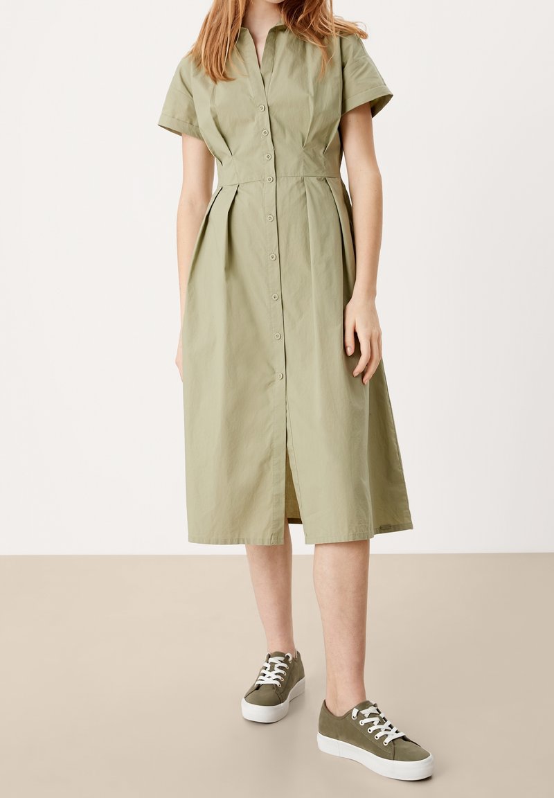 Světle zelené košilové šaty s krátkými rukávy a sklady, s knoflíkovým zapínáním vpředu; v kombinaci s olivově zelenými teniskami a bílými podrážkami.