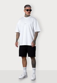White oversized t-shirt with a small logo, paired with black cargo shorts. White sneakers and patterned tattoo on one leg. Dark sunglasses.