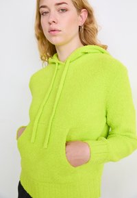 Limeta zeleni dukserica izrađena od teksturiranog materijala, sa prednjim džepom, kapuljačom sa vezicom i rebrastim manžetama i donjim rubom.