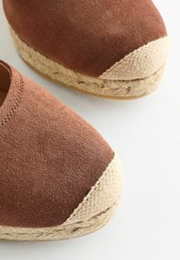 Espadrillas in suede marrone con punta in juta intrecciata di colore crema. Le suole sono leggermente flessibili e hanno una finitura testurizzata.