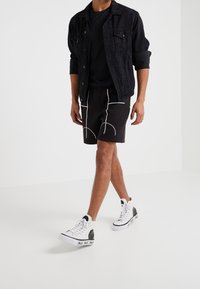 Černá džínová bunda přes černé tričko, spárovaná s černými šortkami s bílými geometrickými vzory a bílými sneakers vysokého střihu s černými akcenty.