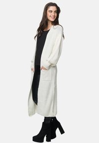 Cardigan lungo a maglia aperta in bianco sporco, con due tasche frontali, spalle ribassate e polsini a costine. Abbinato a una maglietta nera e leggings.