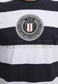 Jūras zilā un baltā svītraina T-kreklu ar īsām piedurknēm, ar apļa logotipu un tekstu "FQ1924 SPORTSWEAR" kontrastējošā gaišā krāsā.