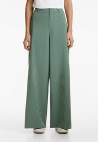 Pantalons large à carreaux verts avec taille haute, présentant une texture lisse et une coupe ajustée, soulignée par un léger pli à l'avant.