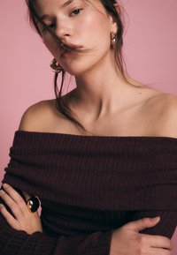 Bordeauxfarbenes, schulterfreies Feinripp-Top mit langen Ärmeln und subtiler Textur, kombiniert mit großen goldenen Creolen vor einem pinken Hintergrund.