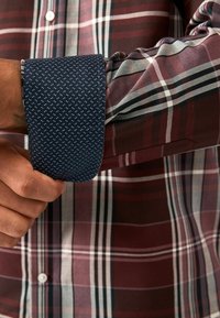 Camisa a cuadros con rayas burdeos y crema, que presenta un puño de lunares azul marino. La tela parece suave con elementos de diseño estructurados.