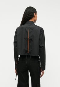 Cămașă neagră scurtă cu decupaj la spate și închidere cu fundiță. Are mâneci lungi și chingi ajustabile pe laterale pentru o potrivire perfectă.