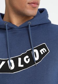 Sudadera azul hecha de una mezcla de algodón con un bolsillo tipo canguro, que presenta un gran logo gráfico en blanco y negro en la parte frontal y detalles de cordones.