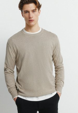 Giovane uomo con pelle chiara e capelli castani, indossa un maglione beige sopra una camicia bianca e pantaloni neri, in piedi con le mani nelle tasche contro una parete bianca.