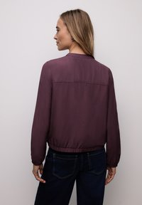 Veste bordeaux à manches longues avec une taille ajustée, un tissu lisse, un col rond et des surpiqûres discrètes le long du dos et des épaules.