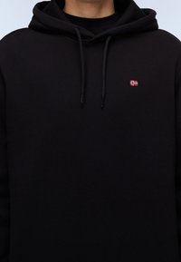 Felpa nera con cappuccio e coulisse, con una piccola bandiera norvegese ricamata sul petto a sinistra, indossata da una persona non visibile.