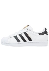 Vita Adidas Superstar-sneaker med svarta ränder, guld "Superstar"-text och snäcka-tådesign, sidovy på vit bakgrund.