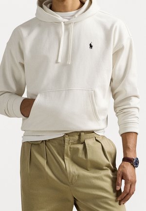 Homme portant un sweat à capuche couleur beige clair avec un logo polo noir, un pantalon plissé kaki, et une montre-bracelet en cuir marron, main dans la poche.