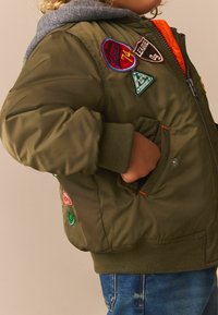 Veste bomber vert olive avec une capuche grise, ornée de plusieurs patchs brodés colorés, des poignets côtelés et une fermeture éclair sur le devant.
