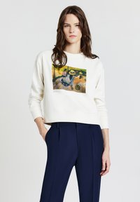 Donna che indossa una felpa bianca con stampa "La Siesta" di Paul Gauguin e pantaloni blu navy, in piedi con una mano in tasca.