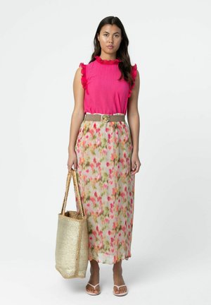 Vrouw die een felroze mouwloze top met ruches draagt, een midi-rok met bloemenprint, een beige riem, witte sandalen en een grote geweven boodschappentas vasthoudt.